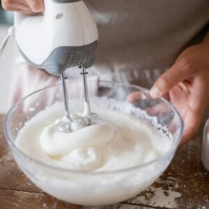Close-up of a person using a hand mixer to make whipped cream in a glass bowl.